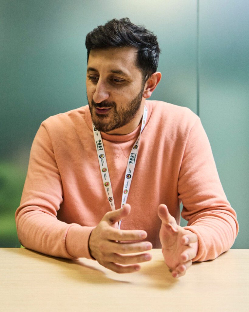 A person sitting at a table, wearing a peach-colored sweater and a lanyard with various symbols. Their hands are gesturing as they speak. The background is blurred.