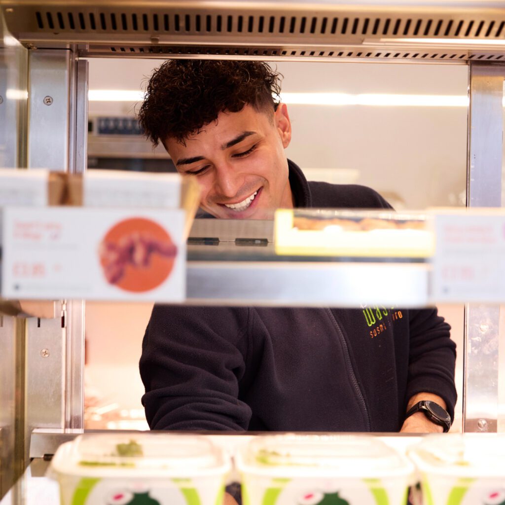 A person smiling while working behind a counter in a shop. He is wearing a dark jacket and appears to be arranging items in a warm display case. Containers of food are visible in the foreground.