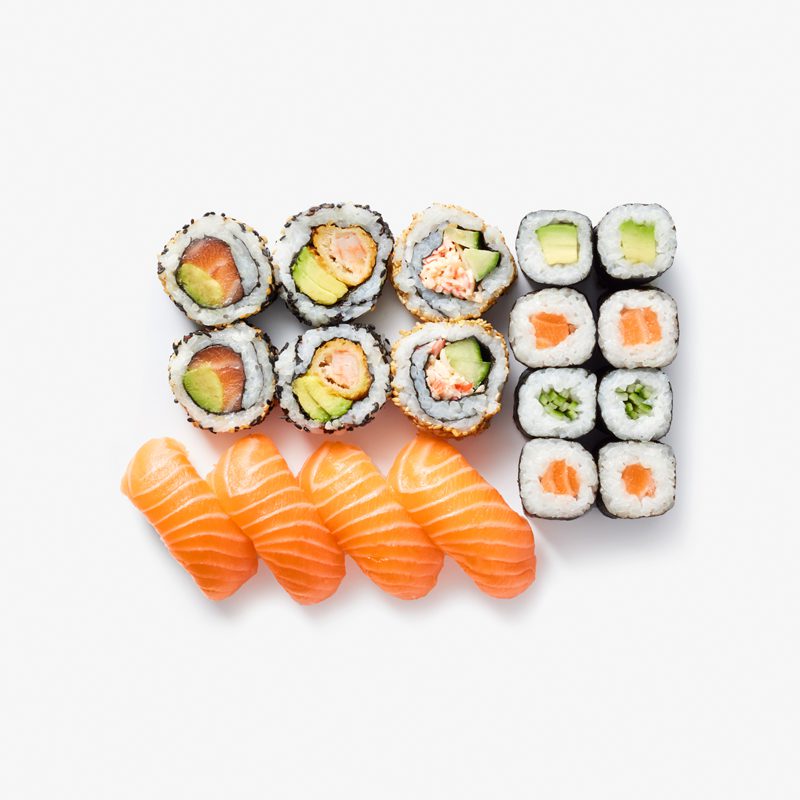 An assortment of sushi displayed in neat rows. Includes salmon nigiri, sushi rolls with avocado and salmon, and California rolls stuffed with crab and avocado. The background is plain white, highlighting the colorful sushi.