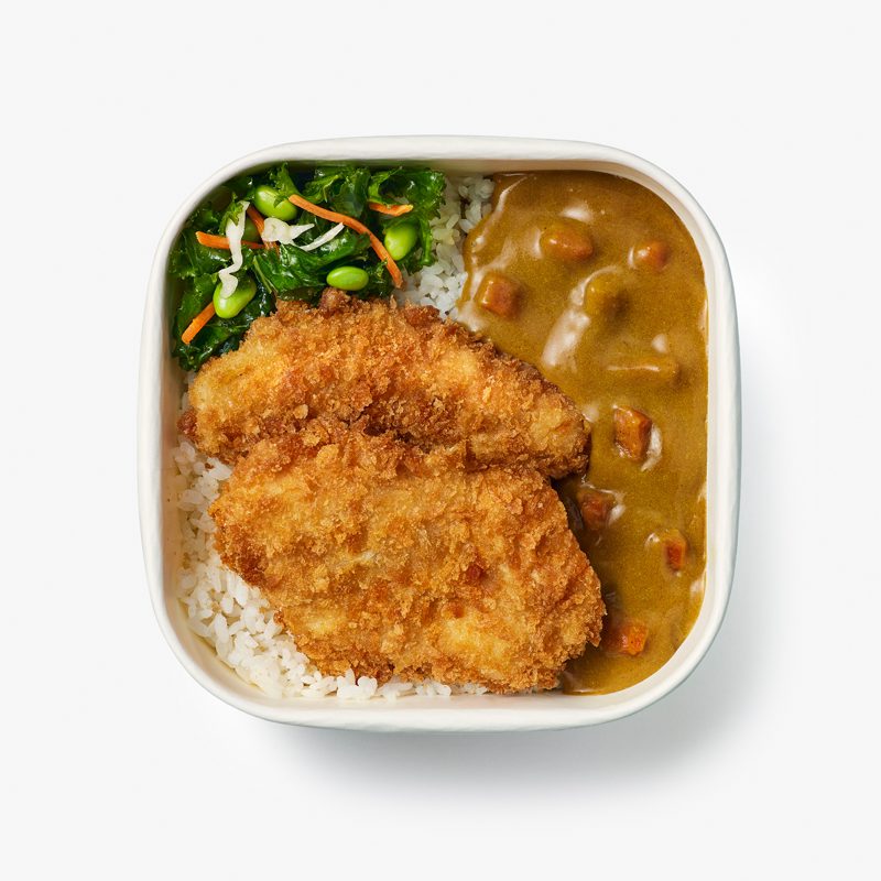 A square dish contains rice topped with two pieces of breaded fried chicken. On the left, there's a serving of green vegetables with carrots. On the right, there's curry sauce with diced vegetables.