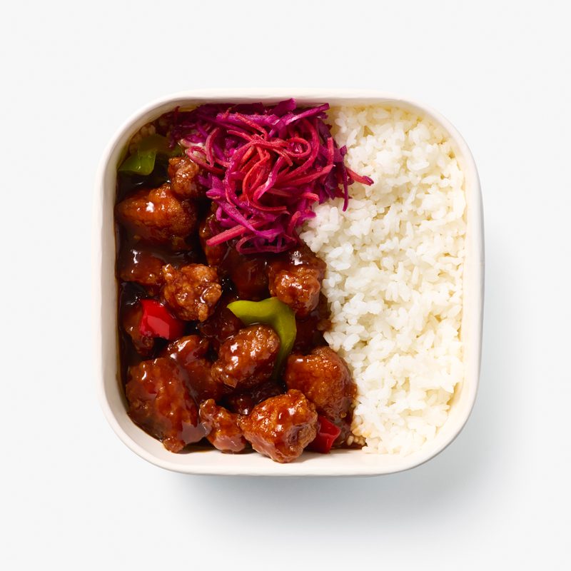 A square dish with white rice, glazed meat chunks with red and green bell peppers, and a garnish of shredded red cabbage on top.