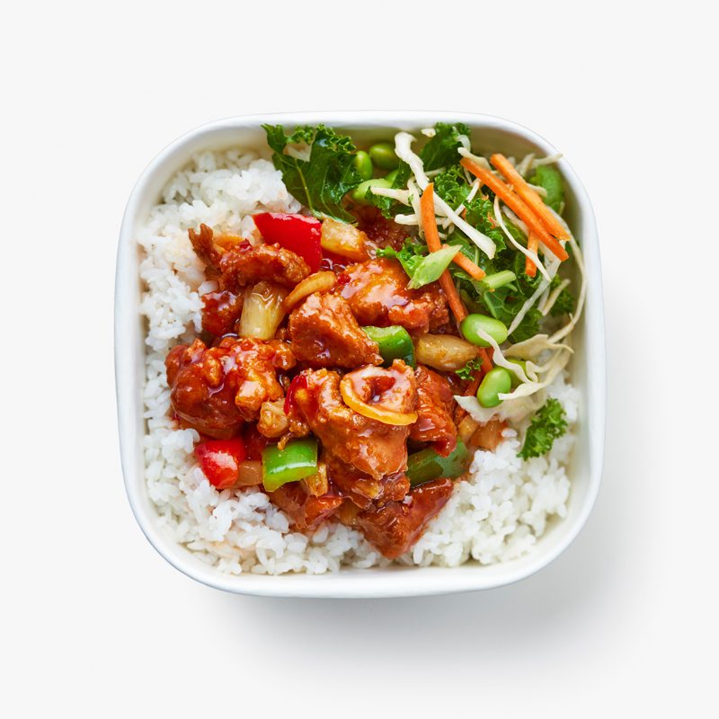 A square white bowl filled with steamed rice, topped with saucy glazed chicken, red and green bell peppers, and garnished with a side of mixed vegetables including shredded carrots, kale, and edamame.