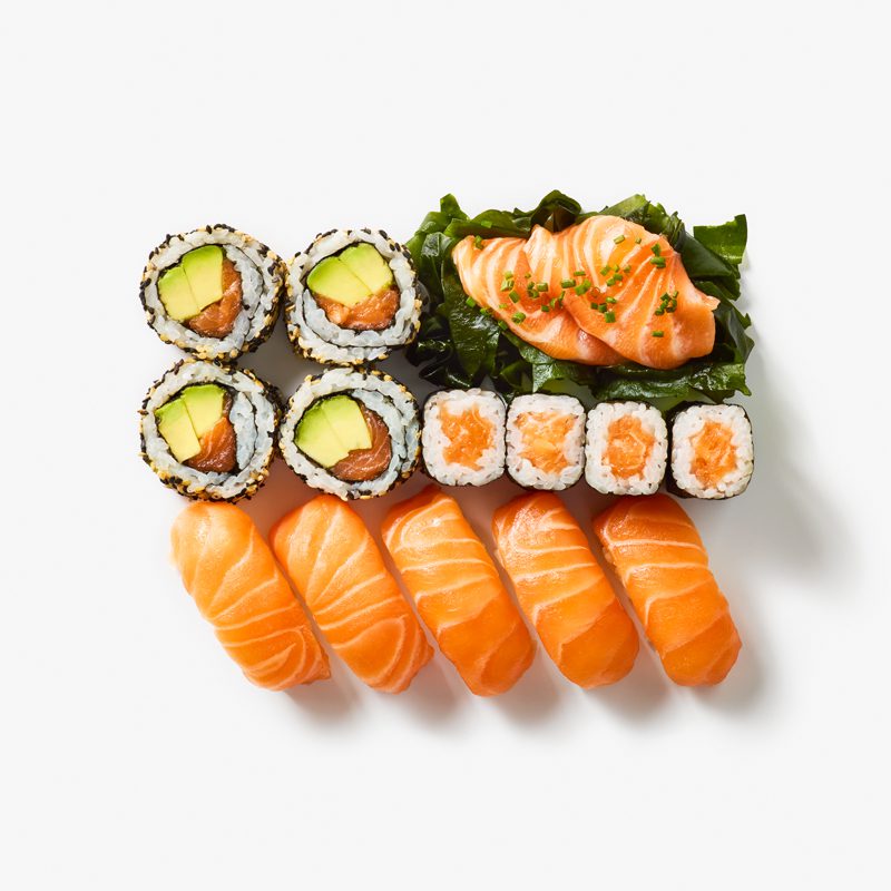 An assortment of sushi on a white background, including salmon sashimi, nigiri, and various rolls with avocado and seaweed. The sushi is neatly arranged in rows, showcasing vibrant green, orange, and black colors.