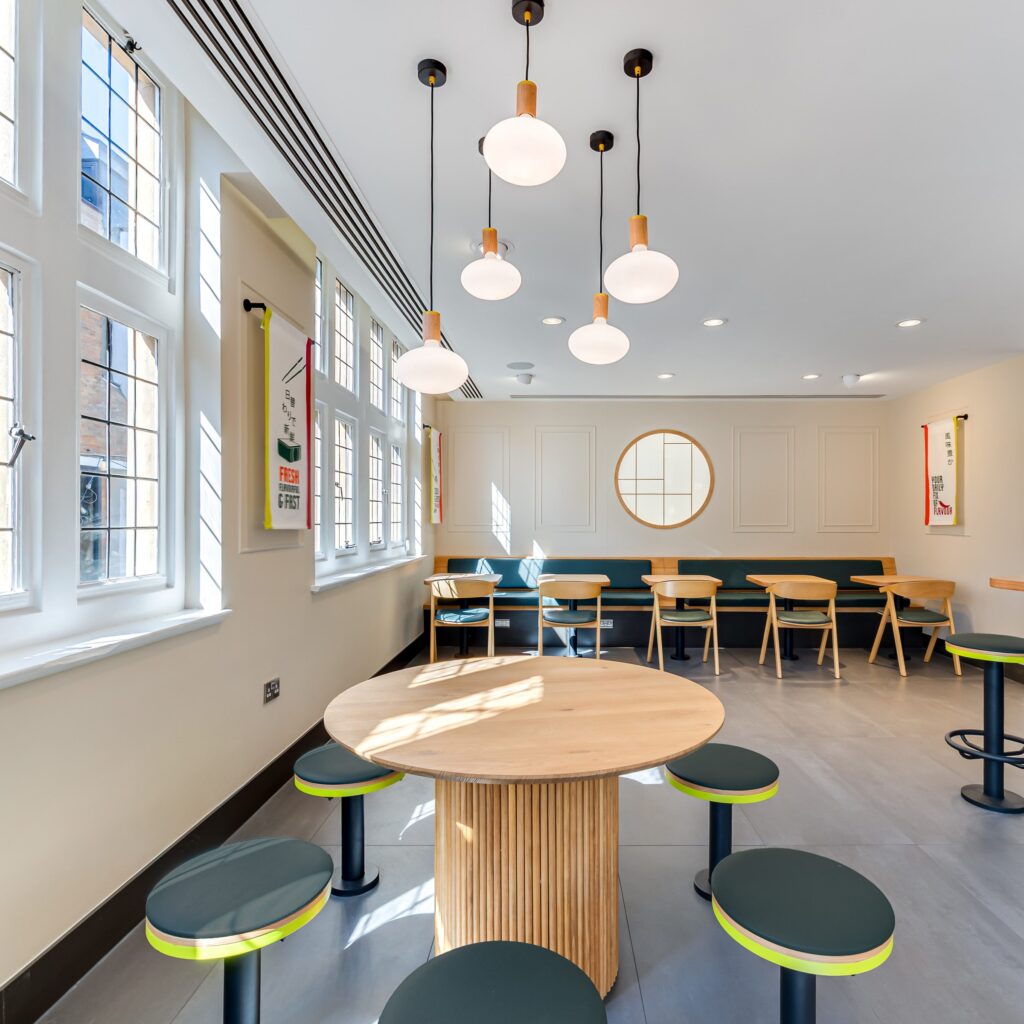 A modern, brightly lit cafe with large windows, round wooden tables, green cushioned stools, wooden chairs along the back wall, pendant globe lights, and a circular mirror on the wall.