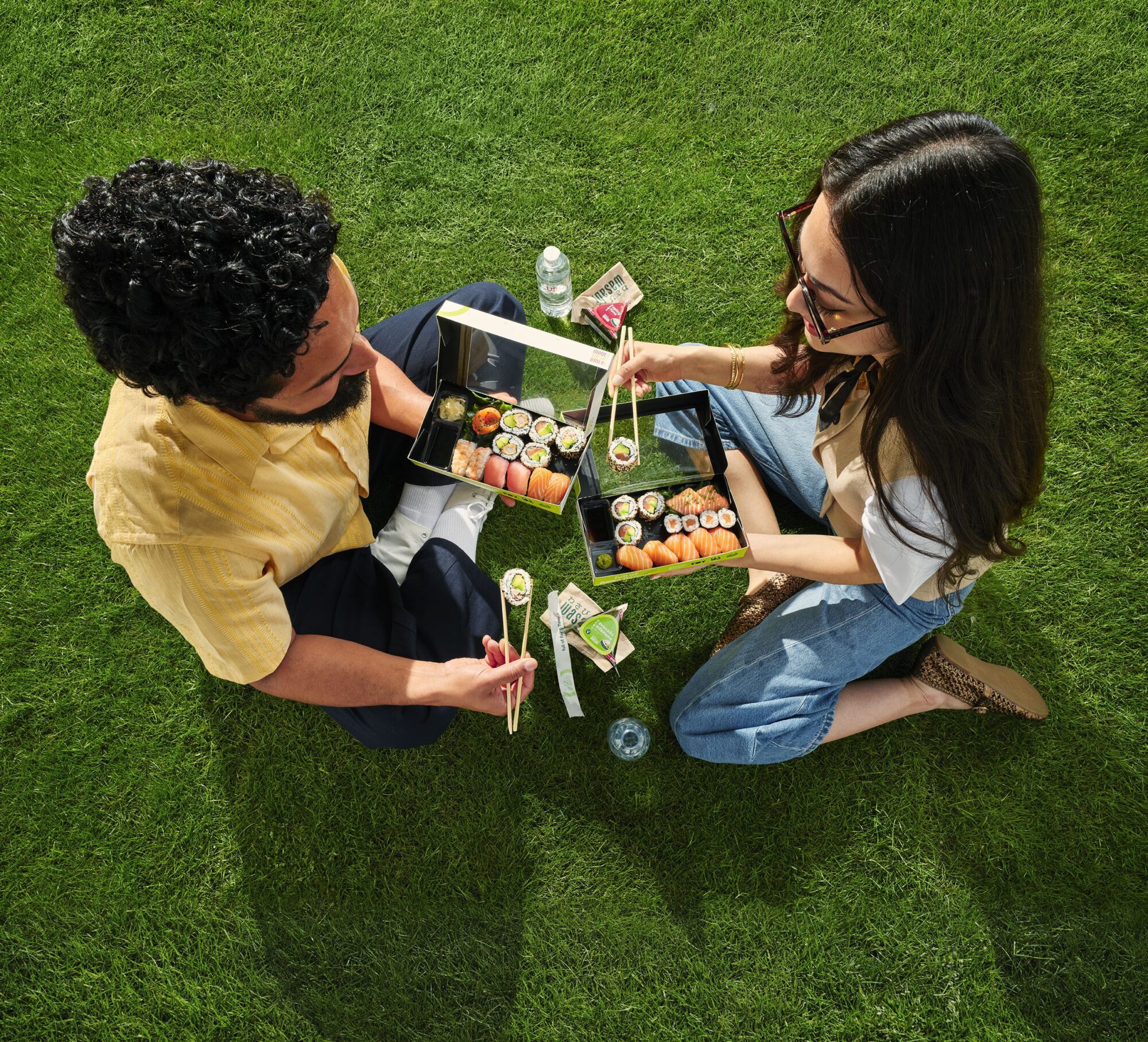 Two people sitting cross-legged on grass, sharing sushi from black trays with chopsticks. They have water bottles, soy sauce, and wasabi sachets nearby. The photo is taken from above.