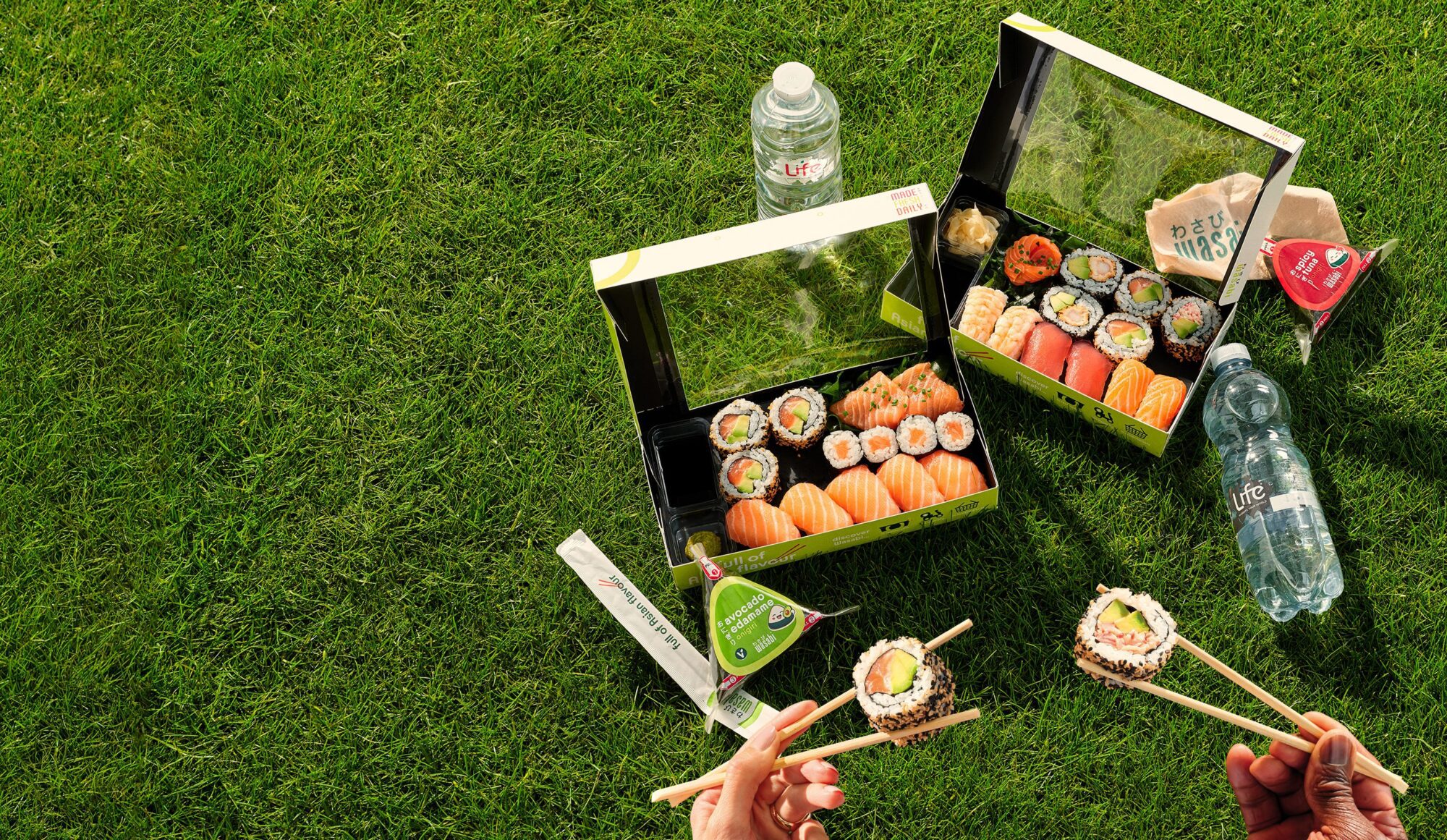Two open sushi boxes, bottled water, soy sauce, and chopsticks are spread out on green grass. Two hands holding sushi pieces with chopsticks are visible in the lower part of the image.