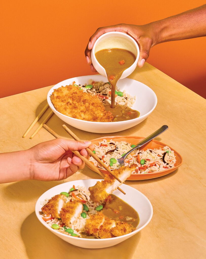 Hands pour curry sauce over a breaded chicken escalope and rice bowl, while another hand uses chopsticks to hold a piece of chicken over a second bowl of curry rice.
